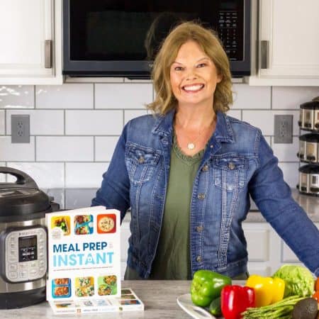 Becca's wearing an olive green dress with a denim jacket and leaning on a white counter. Platter of veggies on the right side of the pic, with an Instant Pot and her book Meal Prep in an Instant on the left side of the pic. Microwave behind her along with white cabinets and subway tile backsplash.