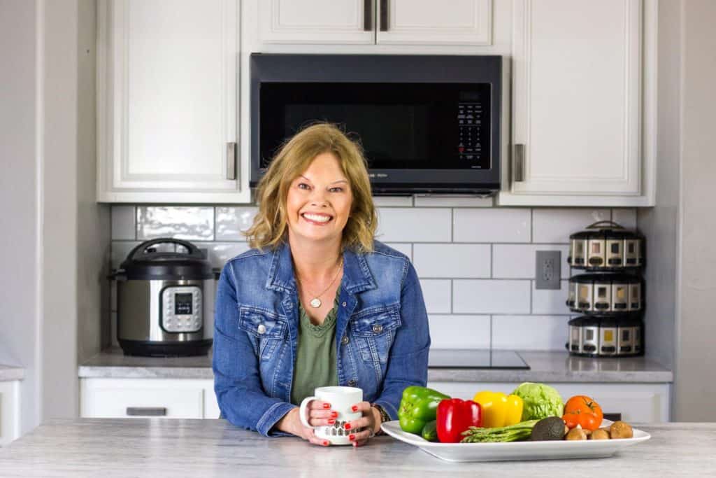 Becca's wearing an olive green dress with a denim jacket and leaning on a white counter holding a light coffee cup. Platter of veggies on the right side of the pic, with a spice rack, Instant Pot, and microwave behind her along with white cabinets and subway tile backsplash.