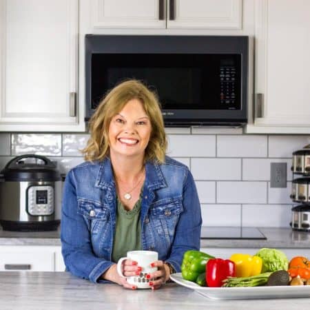 Becca's wearing an olive green dress with a denim jacket and leaning on a white counter holding a light coffee cup. Platter of veggies on the right side of the pic, with a spice rack, Instant Pot, and microwave behind her along with white cabinets and subway tile backsplash.