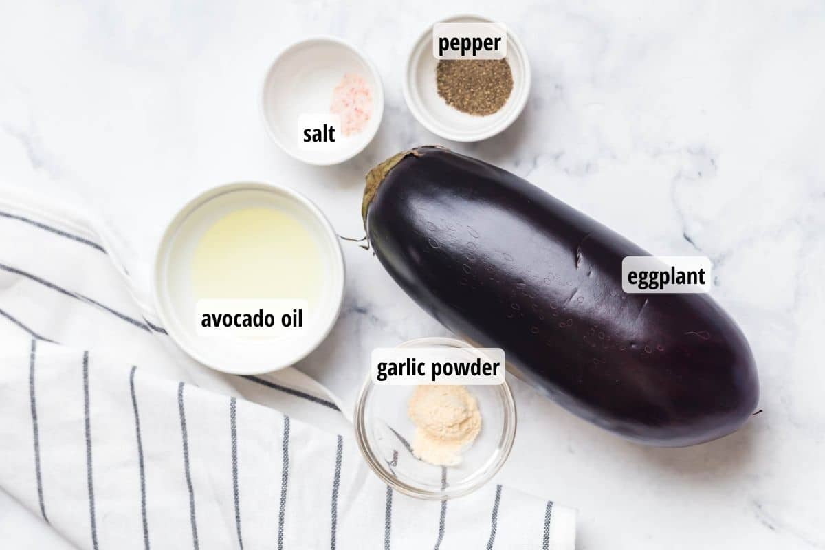 White counter topped with a gray striped white towel, an eggplant, and 4 bowls of spices and oil to make air fryer eggplant.
