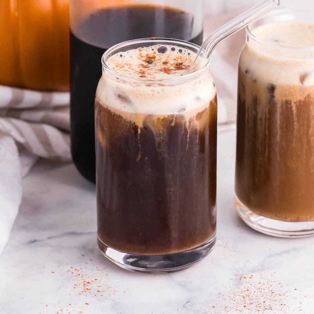 Carafe of cold brew with two clear glasses of pumpkin cream cold brew in front. real pumpkin in the back left of photo. Photo taken on white marble countertops.