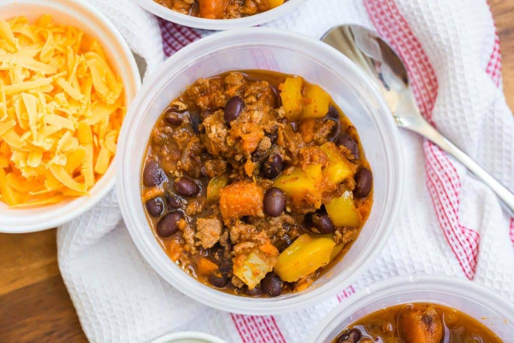 view of sweet potato chili from above on top of a white and red napkin with shredded cheese and a spoon to either side