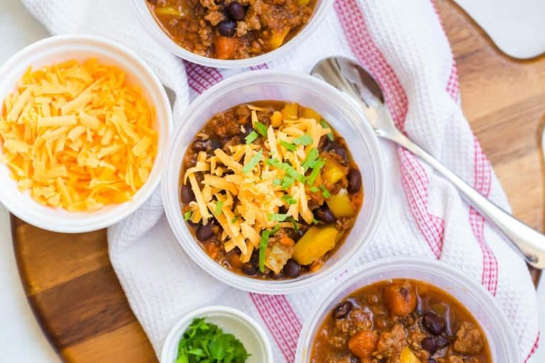 view of sweet potato chili topped with cheese from above on top of a white and red napkin with shredded cheese and a spoon to either side