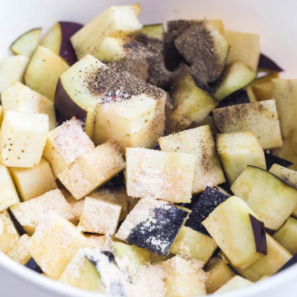 White bowl full of cubed eggplant and topped with seasonings like salt, pepper and garlic powder.
