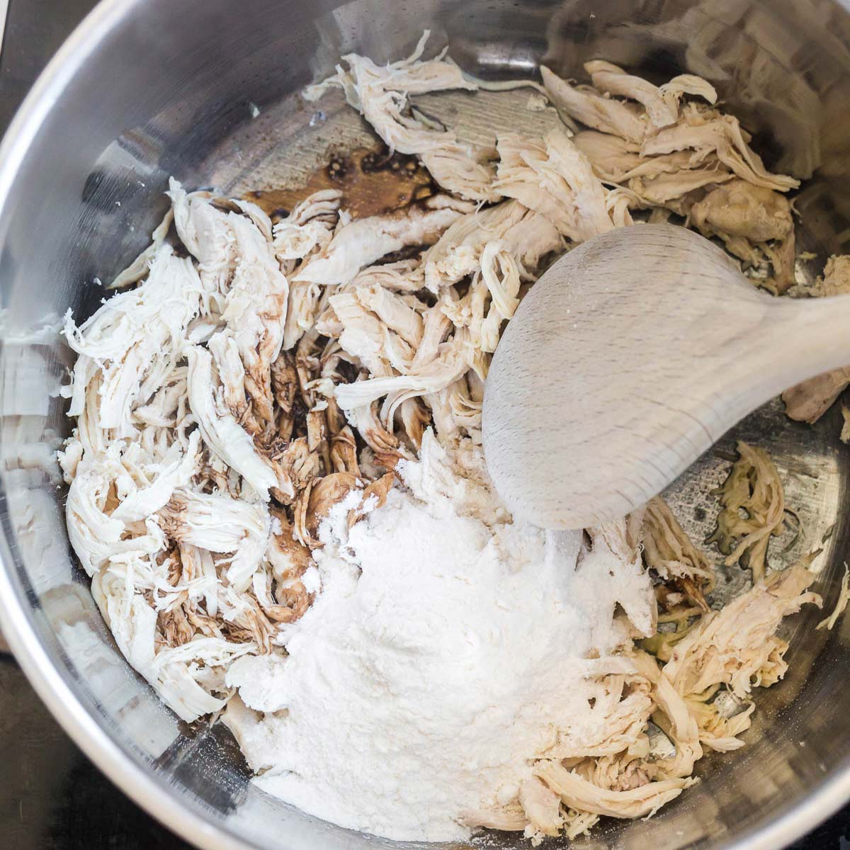 A stainless steel pot with shredded chicken, balsamic vinegar and flour being stirred by a wooded spoon.