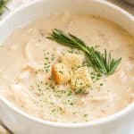 A white bowl with creamy chicken soup topped with a sprig of rosemary, three croutons, and a sprinkling of parsley flakes .
