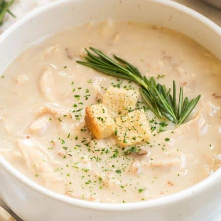 A white bowl with creamy chicken soup topped with a sprig of rosemary, three croutons, and a sprinkling of parsley flakes .