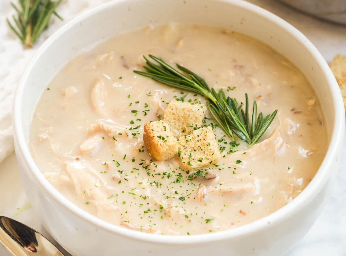 A white bowl with creamy chicken soup topped with a sprig of rosemary, three croutons, and a sprinkling of parsley flakes.