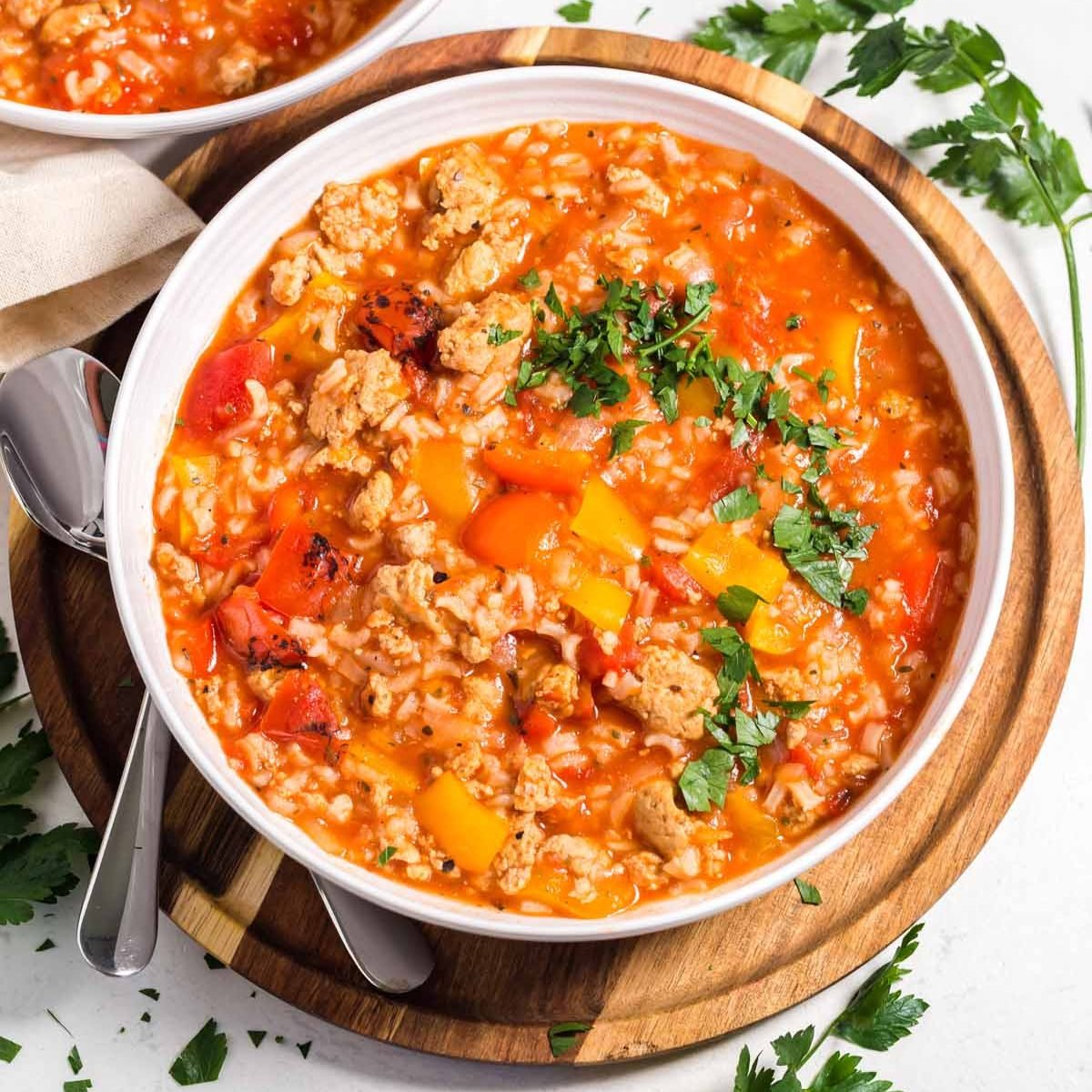 White bowl with stuffed pepper soup on top of a wood platter. There are herbs and another bowl of soup in the background.