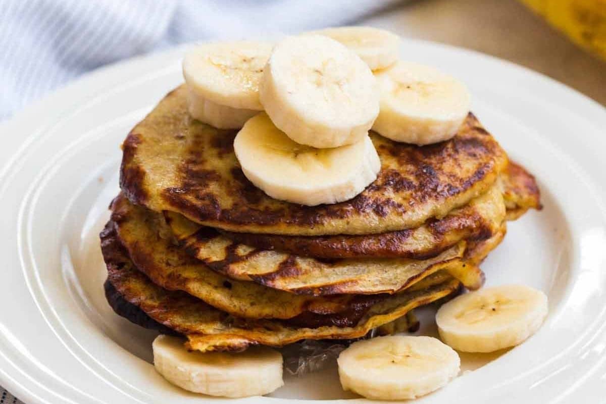 A stack of 2-ingredient banana pancakes on a white plate and topped with slices of bananas.