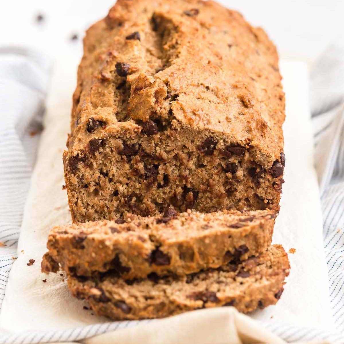 Loaf of banana bread on top of a cream tea towel with a few slices cut off and resting left to the bread.