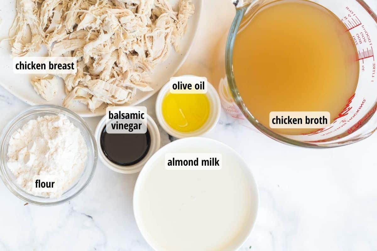 A white counter with labeled ingredients including a plate of shredded chicken, bowls of flour, balsamic vinegar, oil, and almond milk, and a measuring cup of broth.