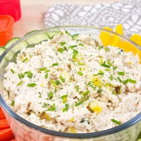 A glass bowl filled with healthy dill pickle chicken salad and surrounded by vegetables like sliced cucumbers, carrot sticks and bell peppers.