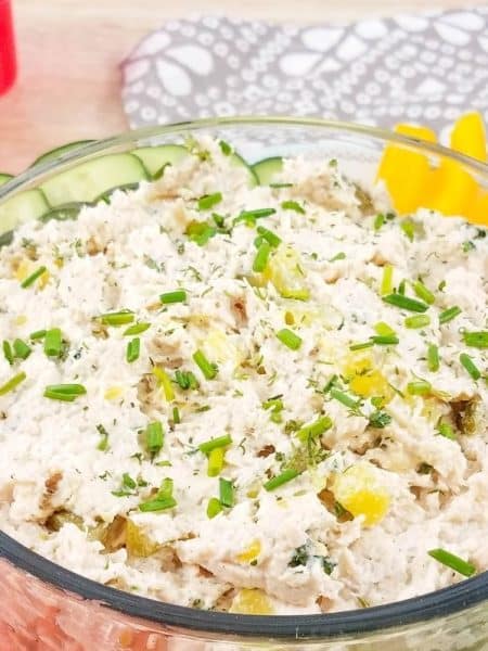 A glass bowl filled with healthy dill pickle chicken salad and surrounded by vegetables like sliced cucumbers, carrot sticks and bell peppers.