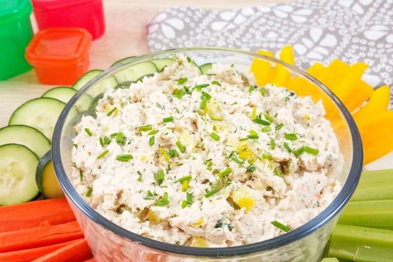A glass bowl filled with healthy dill pickle chicken salad and surrounded by vegetables like sliced cucumbers, carrot sticks and bell peppers.