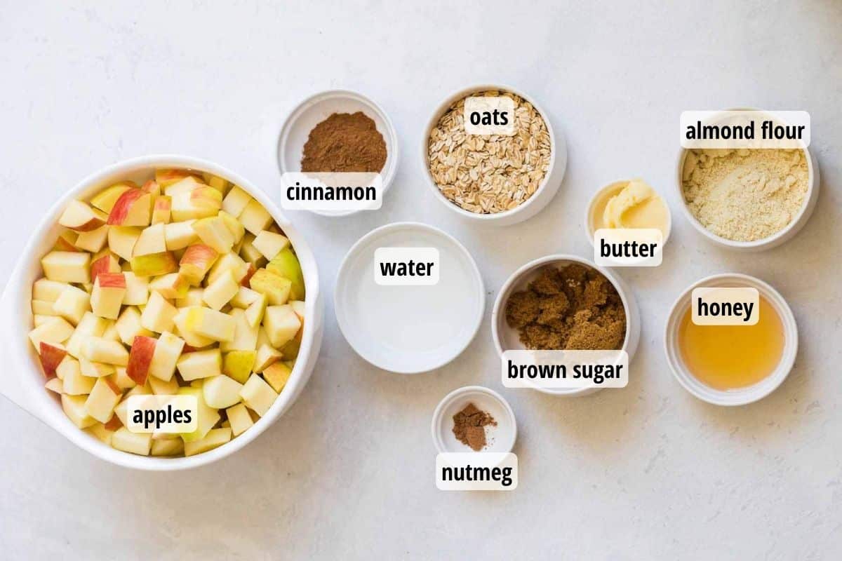 Bowls full of labeled ingredients needed to make healthy apple crumble on top of a marble counter.
