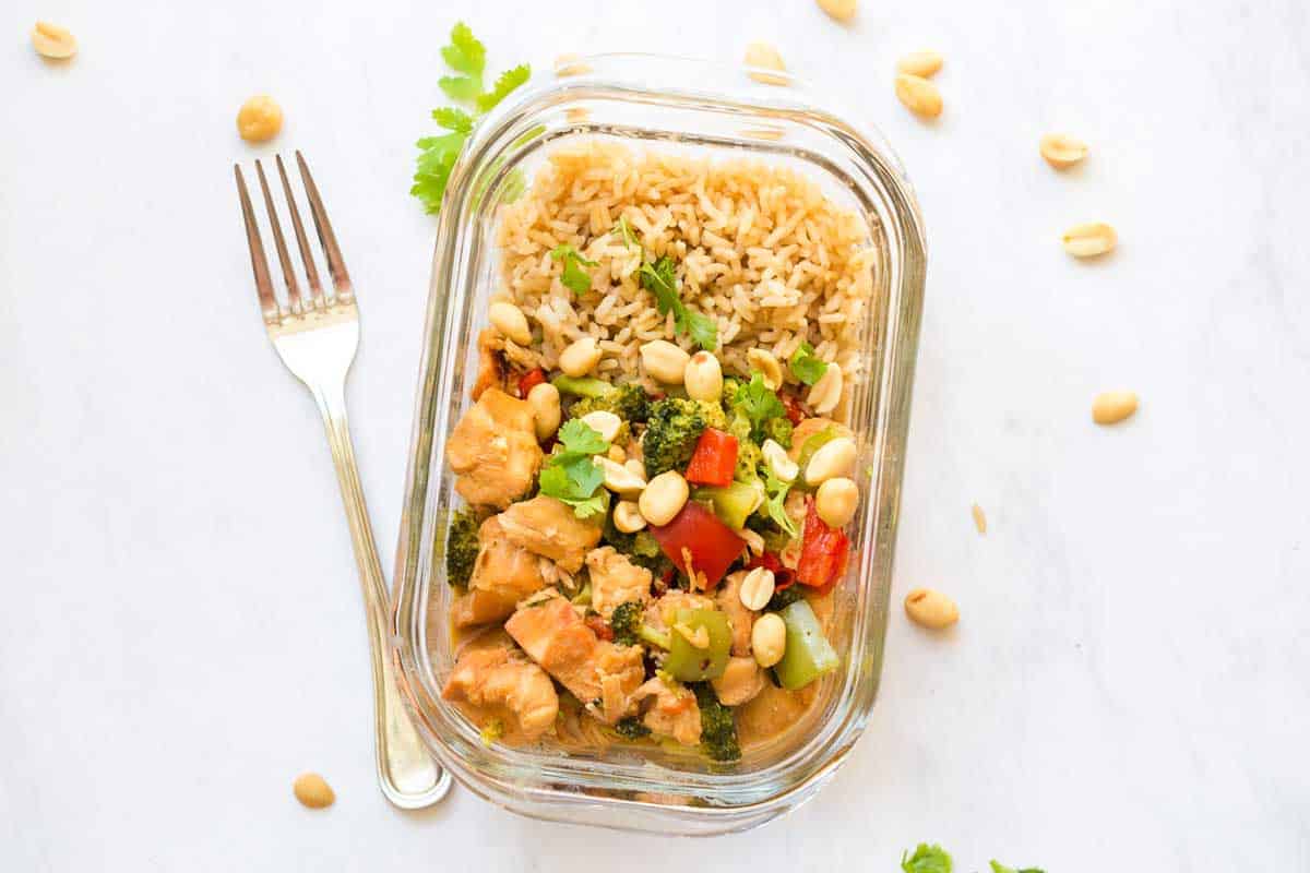 Healthy Kung Pao Chicken in glass meal prep container. There is a fork on the counter next to it, along with parsley springs and peanuts. 