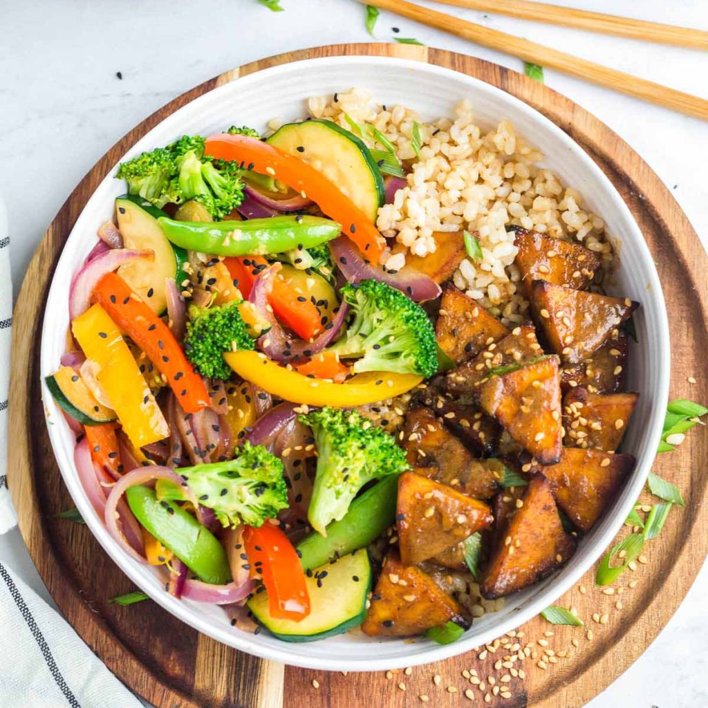 white bowl of teriyaki stir fry with tofu and coconut rice, bowl is on a wooden charger and the chopsticks are above the bowl