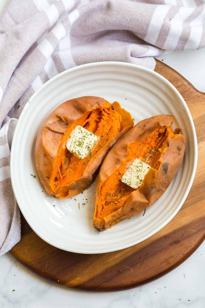 one cooked sweet potato cut in half on on a white plate. slices of butter and chives top each half