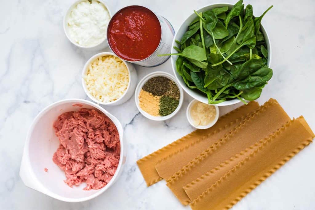 Ingredients for healthy Instant Pot Lasagna sit on a marble countertop in white dishes and bowls. Cottage cheese, cheese blend, seasonings, spinach, ground turkey, and lasagna noodles.
