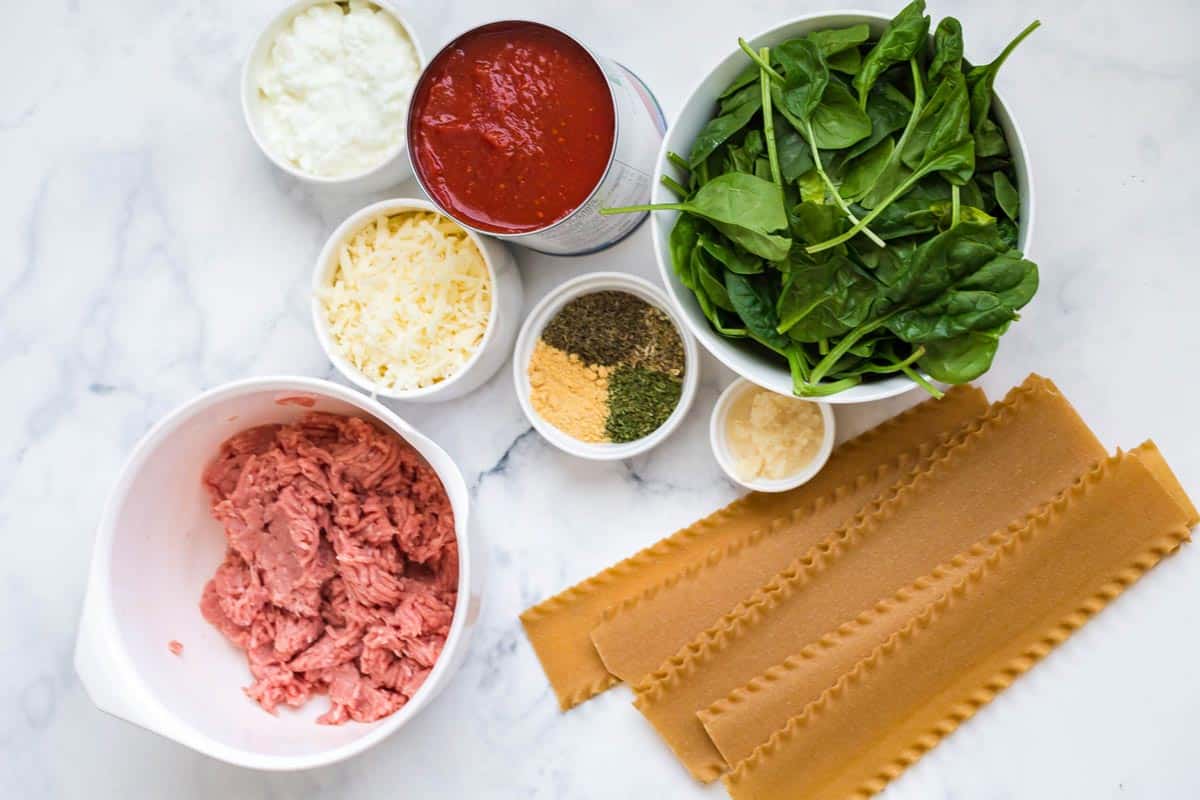 Ingredients for healthy Instant Pot Lasagna on a marble countertop in white dishes and bowls. Cottage cheese, cheese blend, seasonings, spinach, ground turkey, and lasagna noodles.