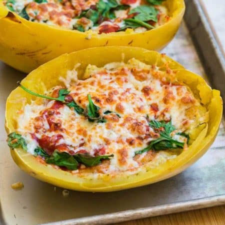 Image of spaghetti squash lasagna halves sitting on metal cooking sheet. The spaghetti squash lasagna is covered with melted and browned cheese with pieces of spinach peeking through the cheese layer.