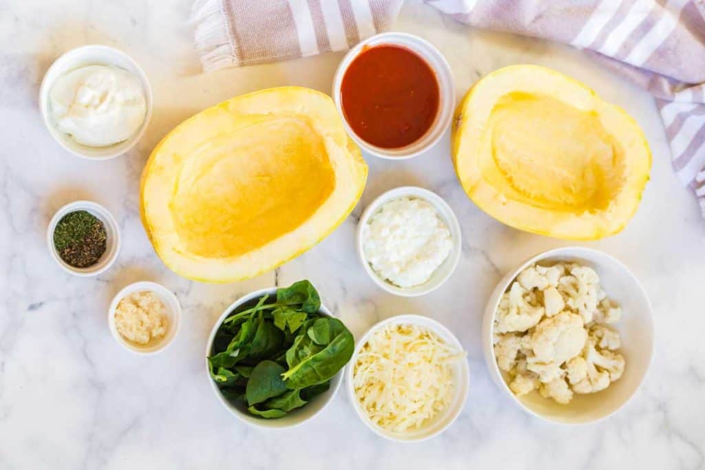 Ingredients of spaghetti squash lasagna are on white countertop. In the image you'll see two seeded and halved spaghetti squash boats. There are also several smaller bowls filled with cheese, spinach, cauliflower, marinara sauce, cottage cheese, greek yogurt, and various spices.