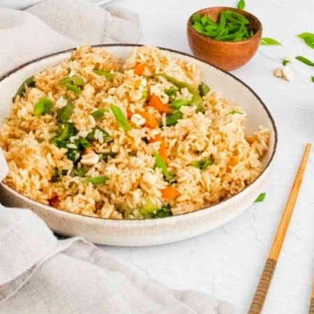 A bowl of homemade fried rice served in a white bowl on a white counter, with small side bowls of chopped green onions and peanuts, and a pair of chopsticks nearby.