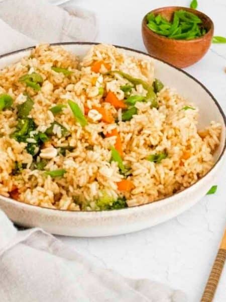 A bowl of homemade fried rice served in a white bowl on a white counter, with small side bowls of chopped green onions and peanuts, and a pair of chopsticks nearby.