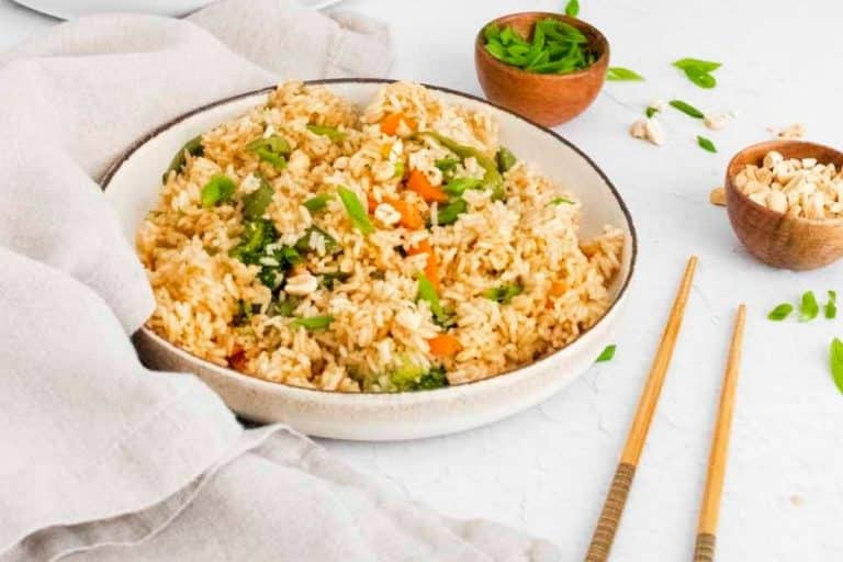 A bowl of homemade fried rice served in a white bowl on a white counter, with small side bowls of chopped green onions and peanuts, and a pair of chopsticks nearby.