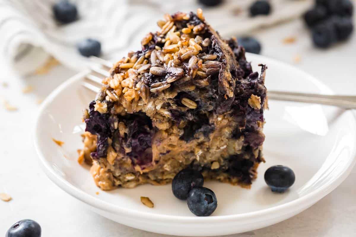 Slice of Blueberry Oatmeal Bake on a white plate with fresh blueberries as a garnish. 