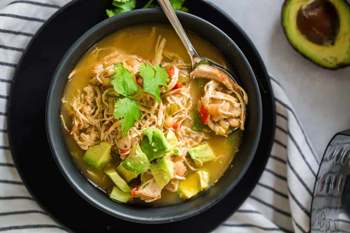 black and white striped napkin with black bowl on top filled with white chicken chili and topped with avocado and cilantro.