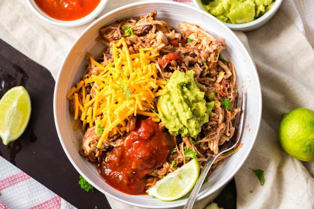 view from above of white bowl with shredded chicken, salsa, guacamole, shredded cheese and a lime wedge
