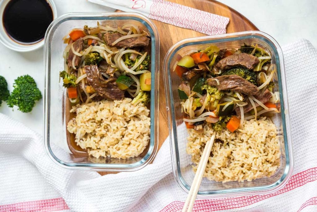 two glass meal prep containers with beef stir fry and cooked brown rice