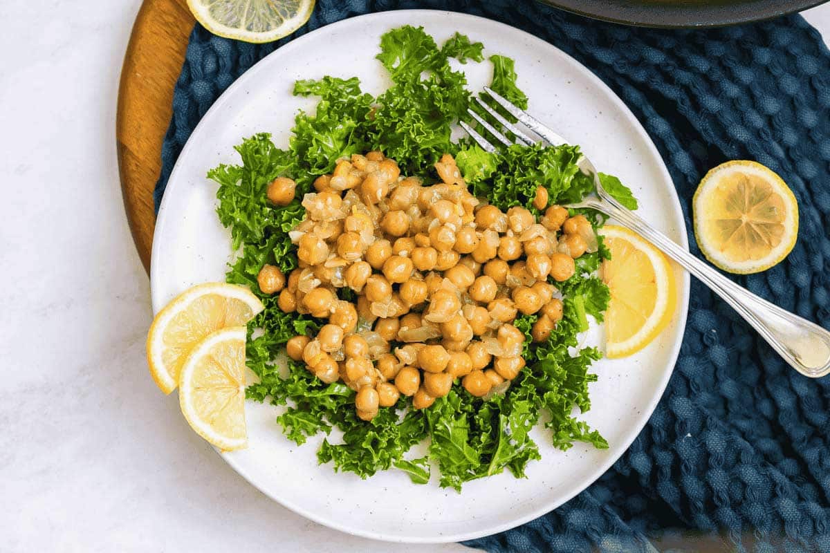 White plate with lemon slices and a bed of kale topped with lemon garlic chickpeas.