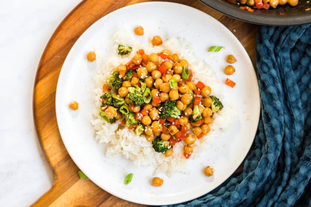 aerial image of white plate, on top of circular wood board, with bed of white rice topped with a chickpea and broccoli mixture