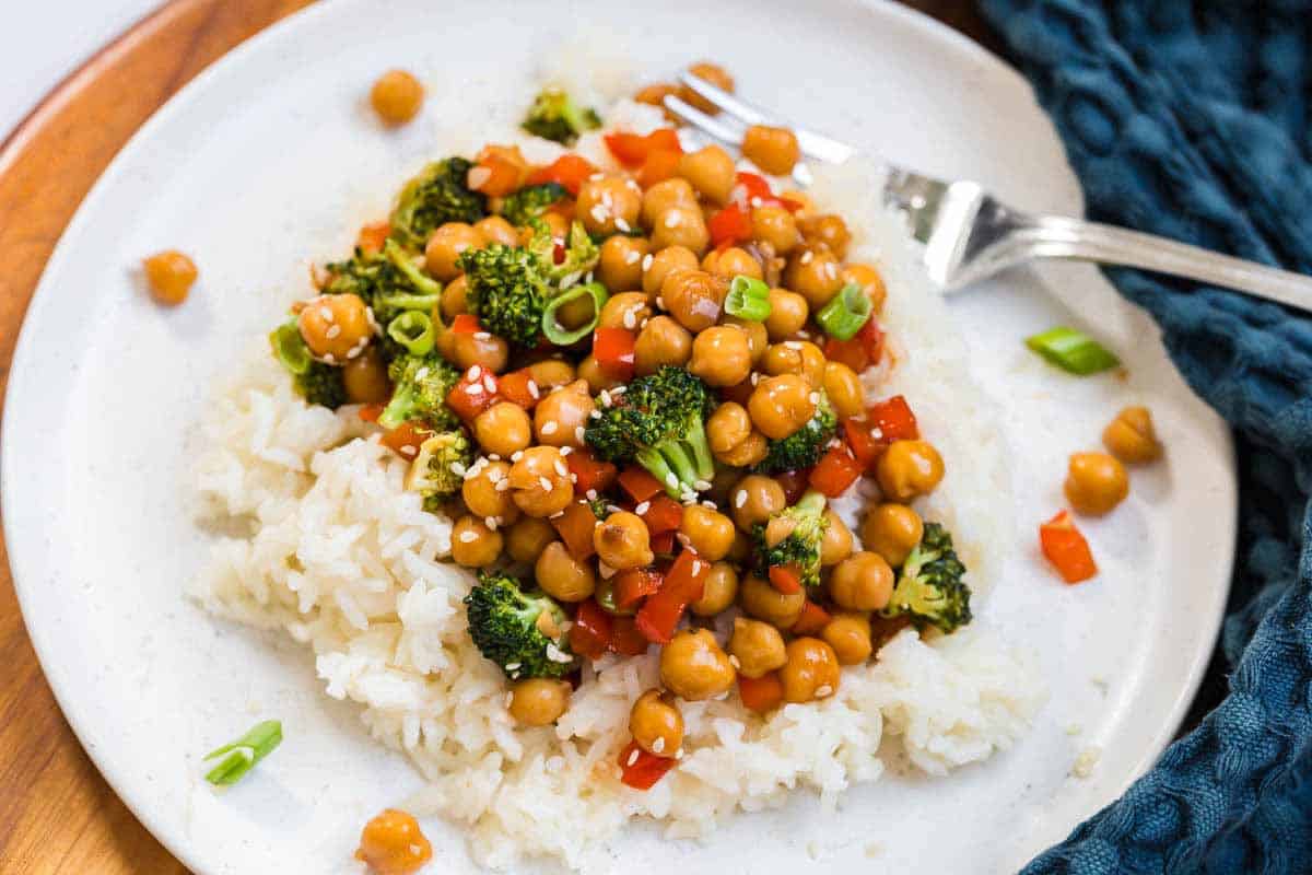 Cooked sweet and sticky sesame chickpeas on a white plate. There is a fork on the right side of the plate and a blue napkin in the background.