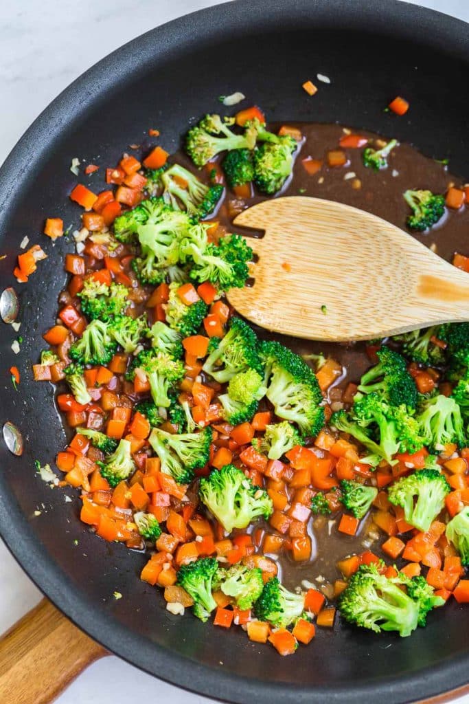 cast iron pan with wooden spoon mixing broccoli, red pepper, and spices.