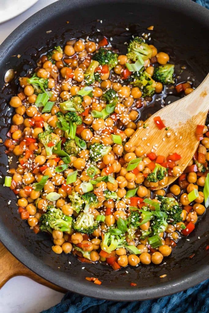 cast iron pan with wooden spoon mixing broccoli, red pepper, and chickpeas. Toppings include diced green onions and sesame seeds. 