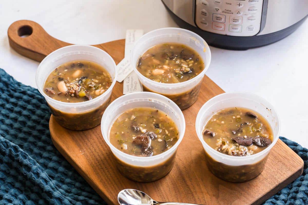 Four meal prep bowls filled with mushroom soup sitting on top of a wooden board which is on top of a blue kitchen towel.