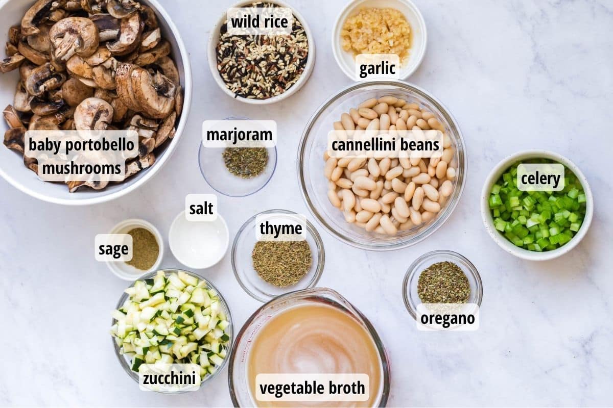 A white marbled counter top with bowls full of 12 different labeled ingredients needed to make wild rice and mushroom soup.
