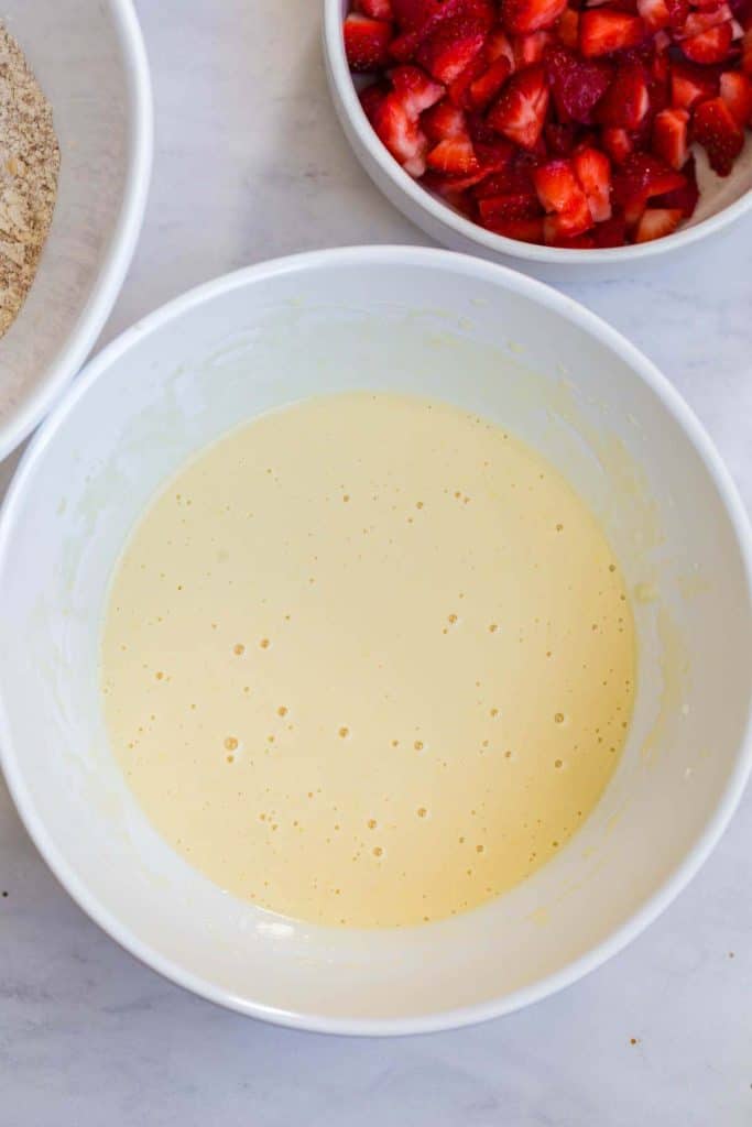 white bowl on white countertop with wet batter in it, white bowl of cut strawberries at the top right
