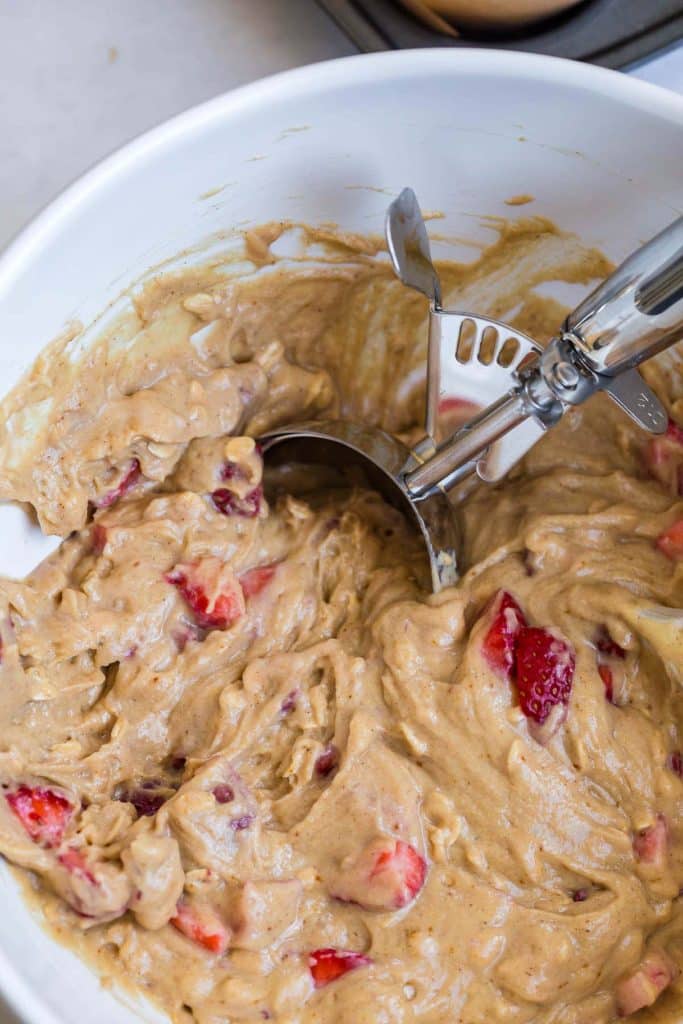 image of cut strawberries mixed in with muffin batter, cookie scoop is in the bowl as well