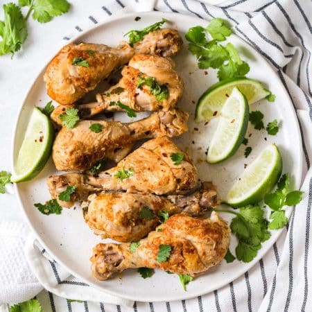 a white and gray plate with cilantro lime chicken drumsticks on it. Garnished with pieces of cilantro and lime wedges. Plate is on top of a black and white striped napkin.