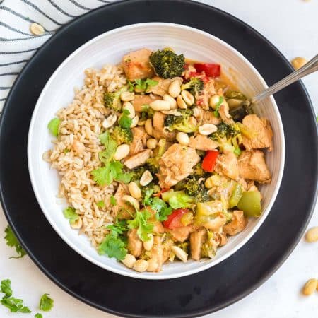 top down view of white bowl on a black dinner plate. The bowl contains cooked Kung Pao chicken with peanuts, broccoli, and peppers, and there is also brown rice in the bowl.