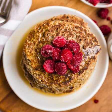 a white plate of vegan pancakes with raspberries on top, drizzled with syrup. In the background you can see a part of a fork and some more raspberries.