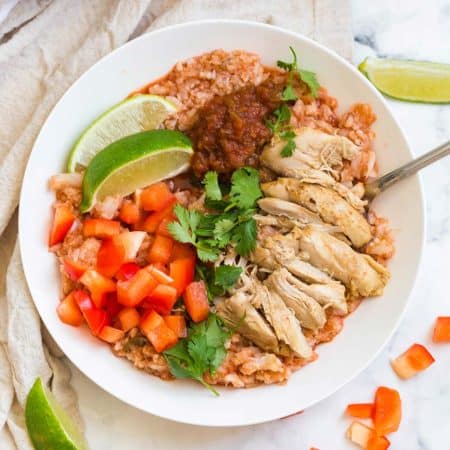 top down view of white bowl on a white counter with cooked cauliflower rice, chicken thighs, red peppers, cilantro, and salsa on top.