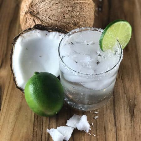 close up of a clear glass with ice and a margarita in it. THere is lime garnish on the top and the cocktail is sprinkled with shredded coconut. There is a lime off to the left side and a coconut, cut in half, behind the glass.