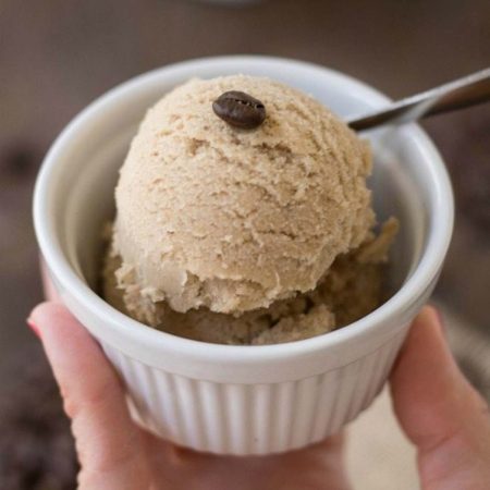 close up of a scoop of dairy free coffee ice cream with one espresso bean on top. Ice cream is in a white dish being help by a woman's hand