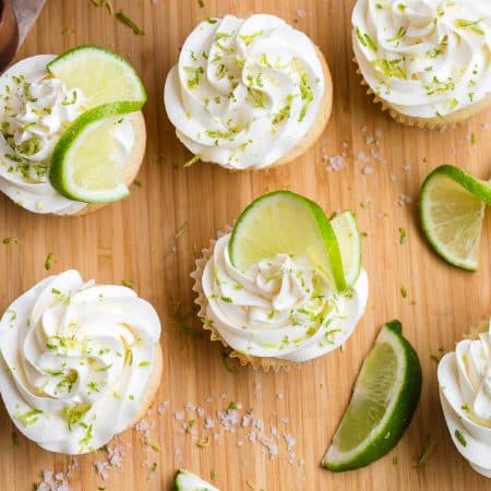 top down image of 6 margarita cupcakes garnished with lime zest and lime twists. The cupcakes are sitting on a wood cutting board, and have coarse salt sprinkled next to lime wedges.